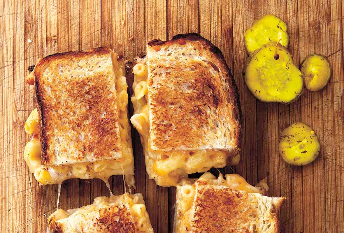 A grilled mac and cheese sandwich cut into four squares on a wooden board with pickle slices beside it.