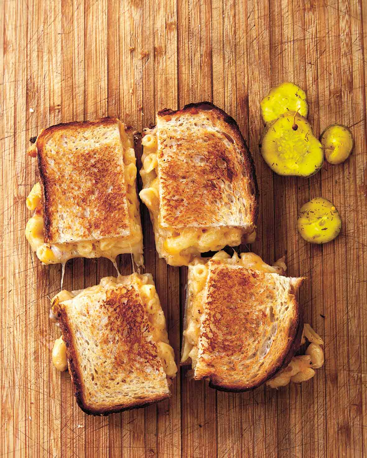 A grilled mac and cheese sandwich cut into four squares on a wooden board with pickle slices beside it.