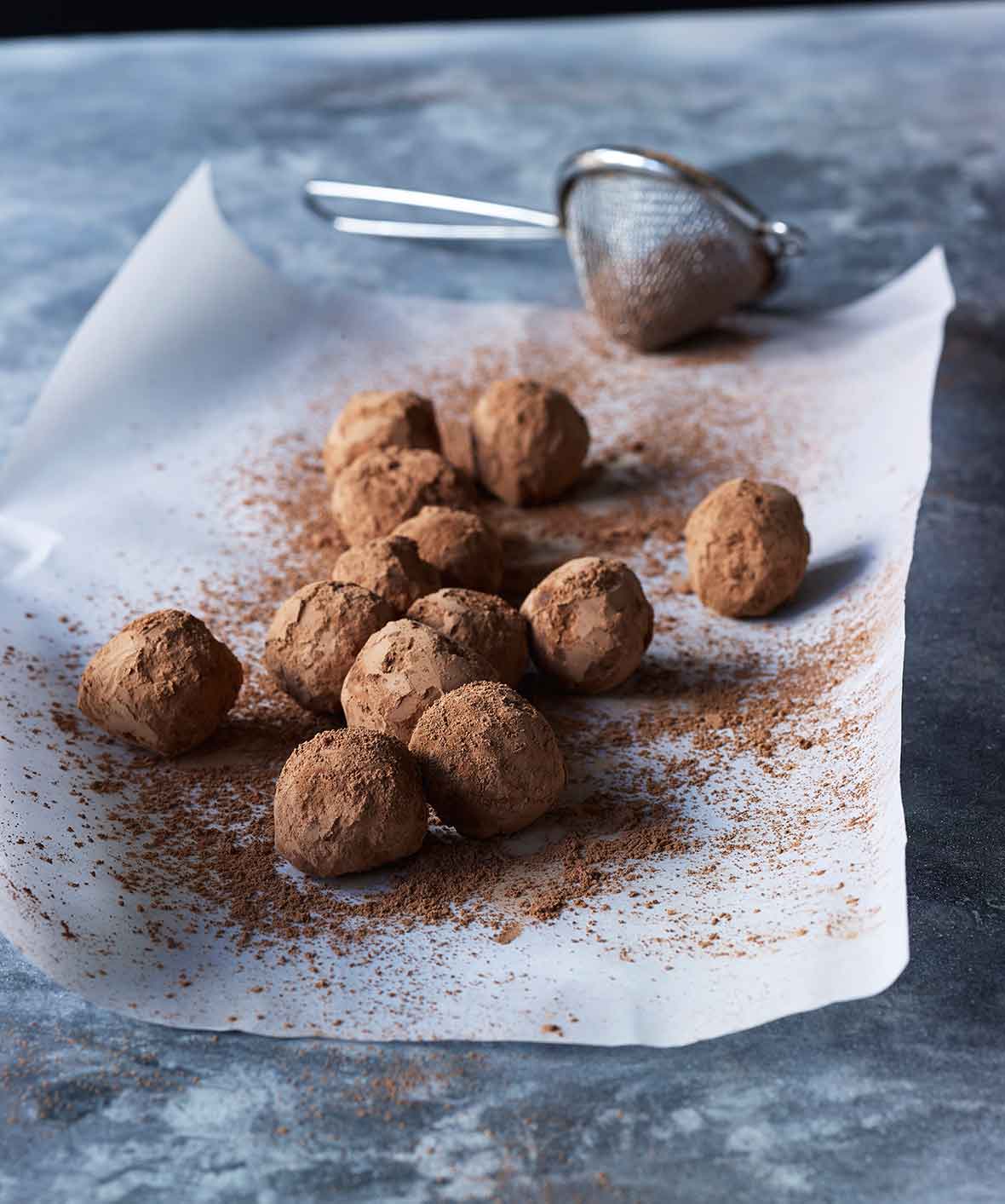 A sheet of parchment paper topped with chocolate truffles and a sifter of cocoa powder on the side
