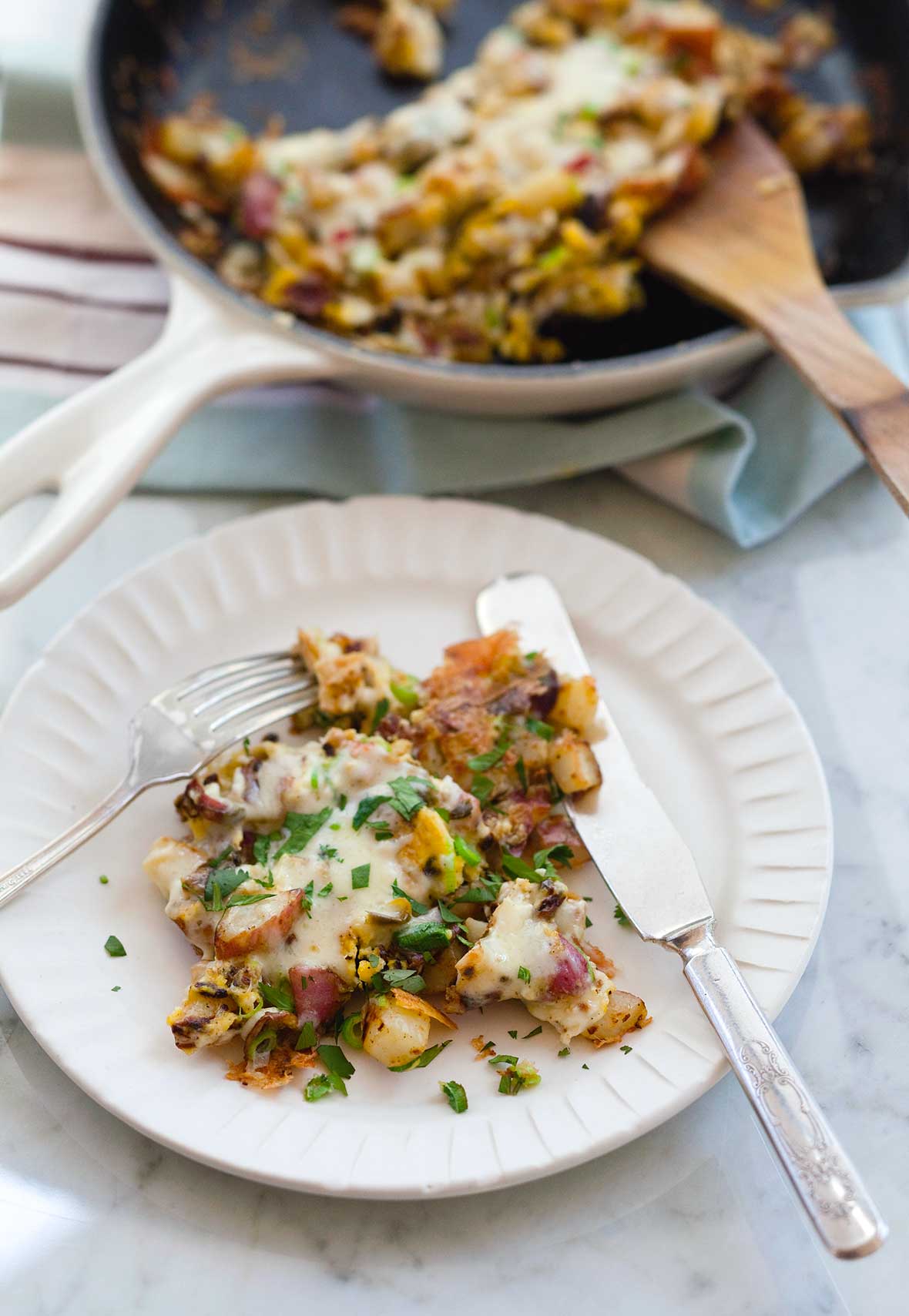 A white plate of fried potatoes with eggs, covered in cheese with a skillet in the background and a fork and knife on the plate.