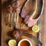A cutting board with lamb chops, some in racks, some sliced, along jus and lemon halves
