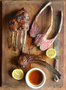 A cutting board with lamb chops, some in racks, some sliced, along jus and lemon halves.