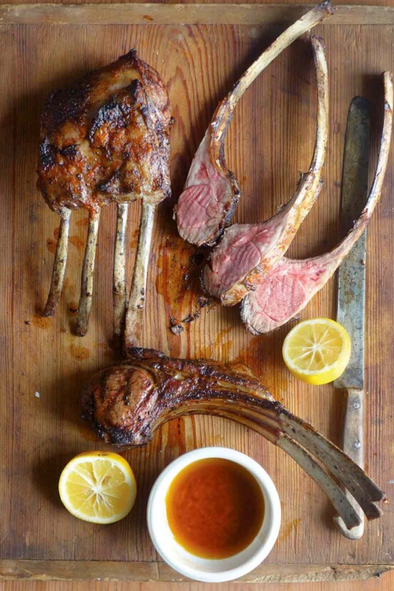 A cutting board with lamb chops, some in racks, some sliced, along jus and lemon halves.