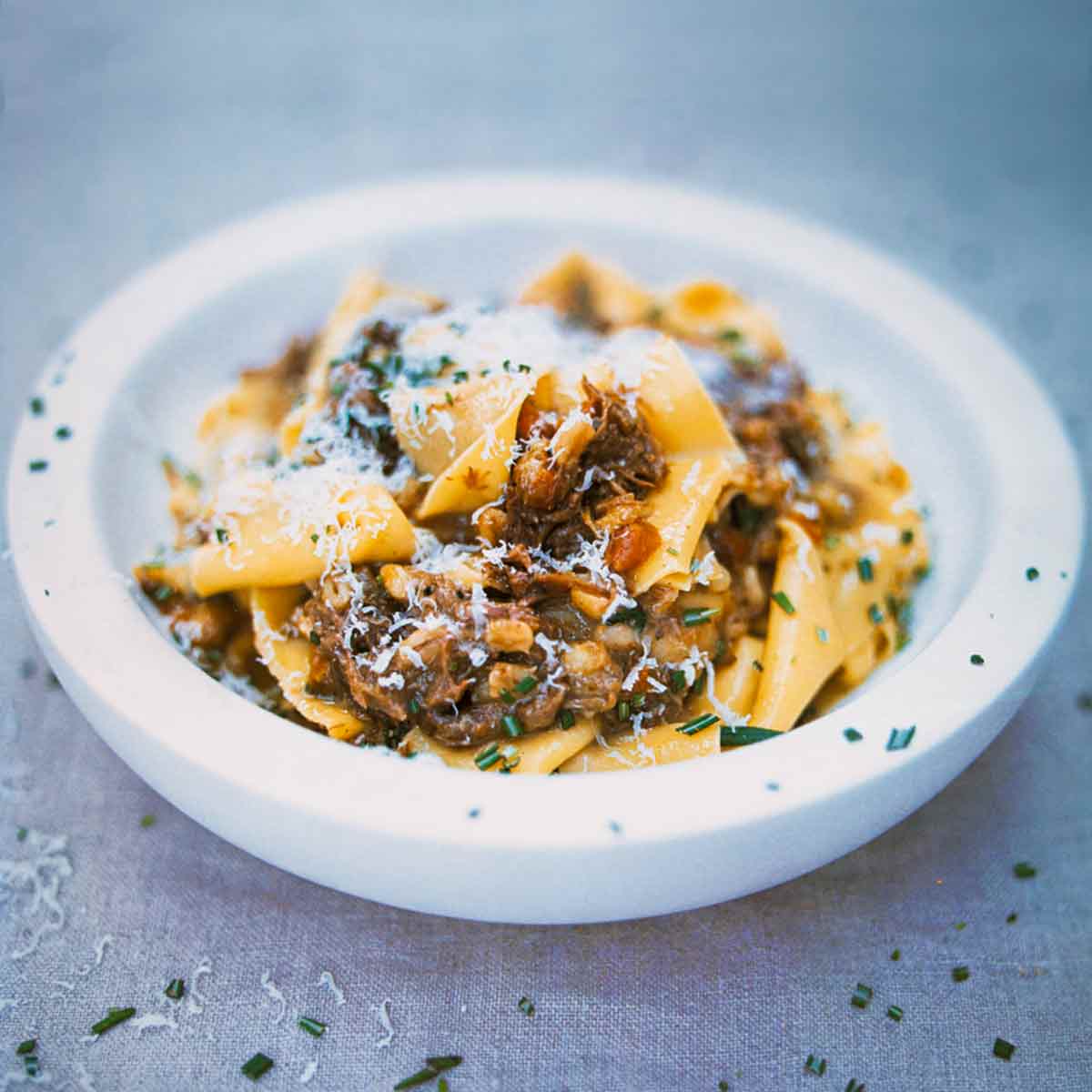 A white bowl filled with pappardelle pasta with meat sauce sprinkled with chopped rosemary.