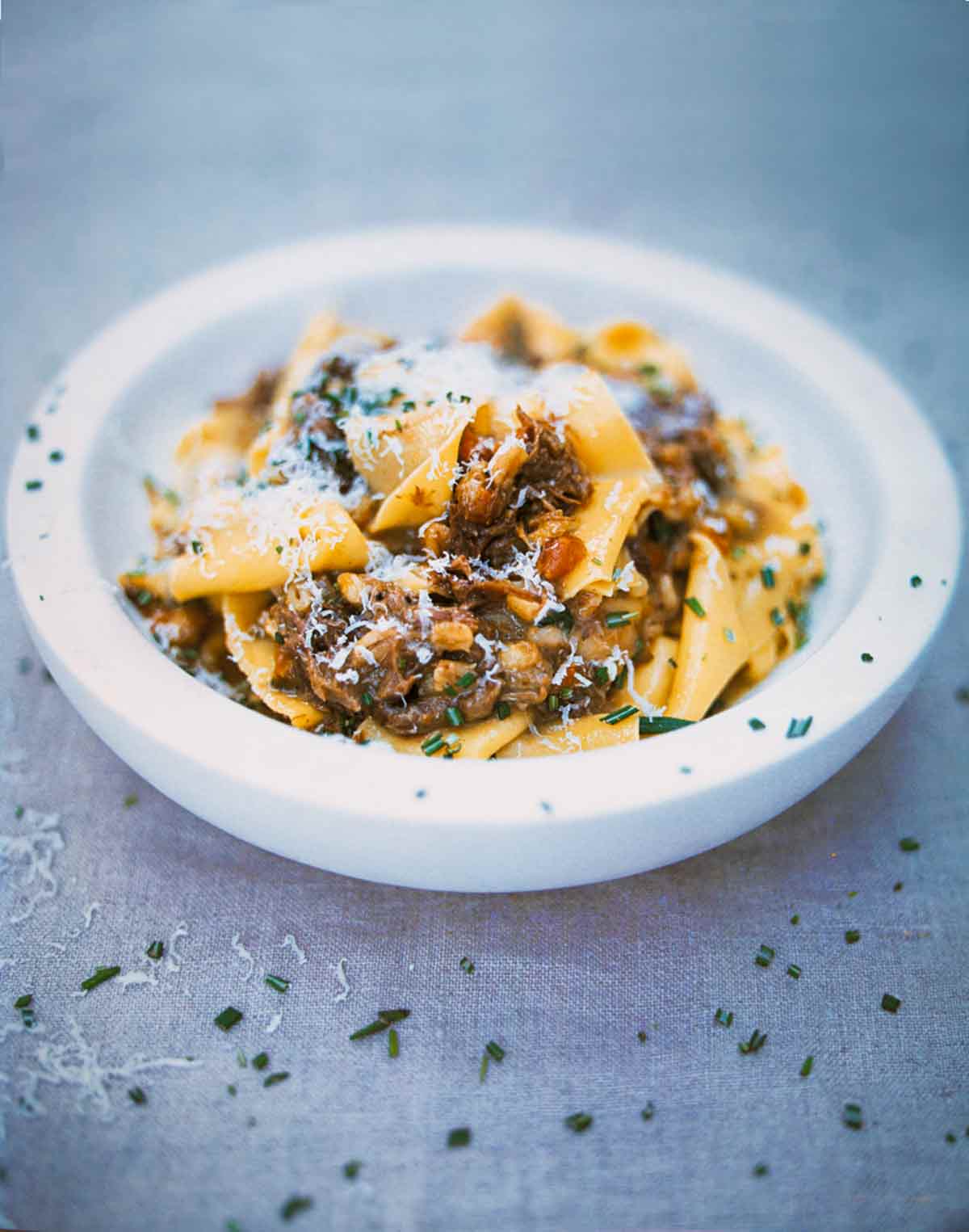 A white bowl filled with pappardelle pasta with meat sauce sprinkled with chopped rosemary.