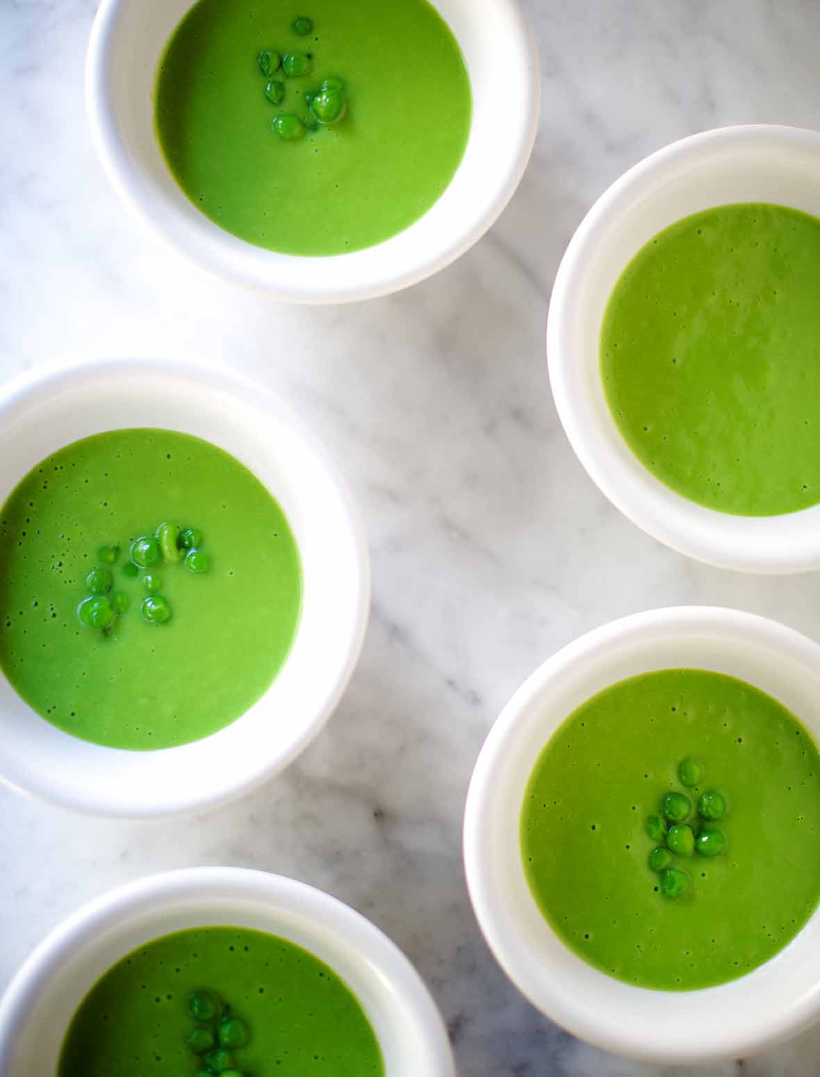 Five white bowls filled with pureed cold sweet pea soup, topped with a few who peas