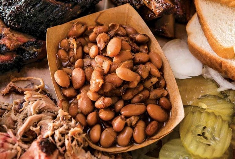 A paper contain of barbecue beans and pulled pork, surrounded by hunks of smoked meats