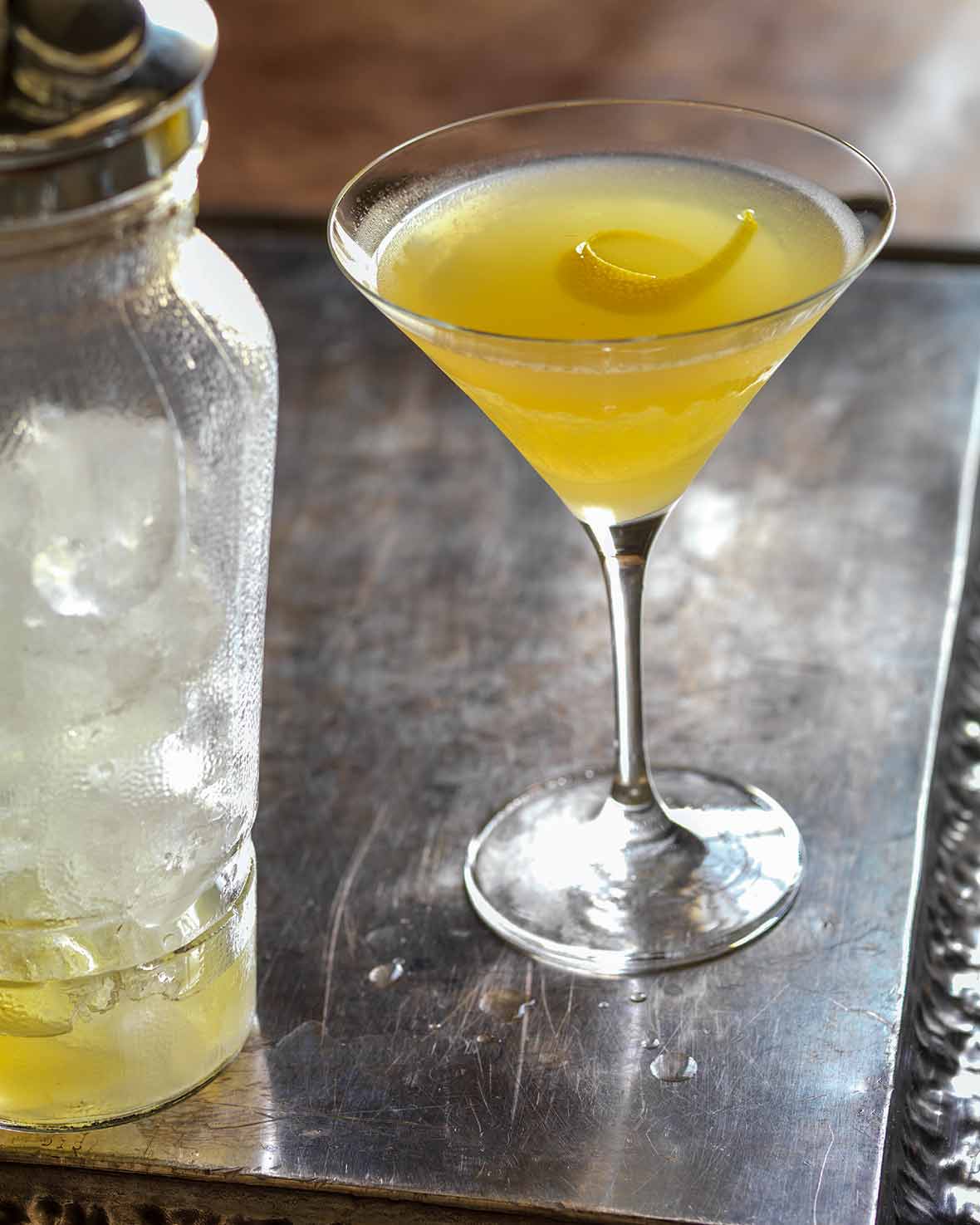 Martini glass filled with yellow Bee's Knees cocktail--honey, gin, and lemon--with a lemon twist, a shaker on the side