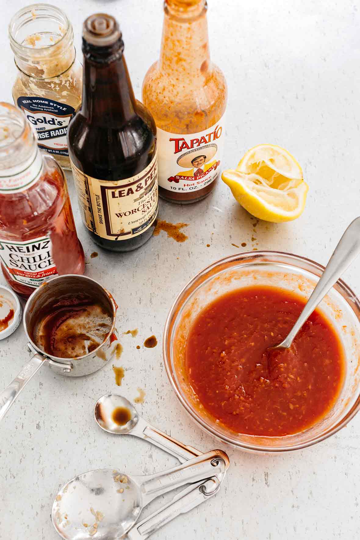 A glass bowl of homemade cocktail sauce with a spoon resting inside and bottles of chili sauce, hot sauce, and Worcestershire sauce in the background.