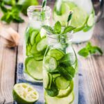 Two small glass bottles filled with cucumber water, cucumber slices, lime slices and mint on a wooden table.