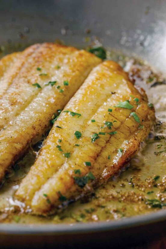 Two fillets of flounder in a lemon butter sauce in a skillet topped with chopped parsley