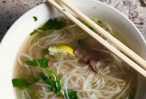 A bowl of authentic Vietnamese pho with rice noodles, beef, lemon, basil, and scallions in it and a pair of chopsticks resting on top of the bowl.