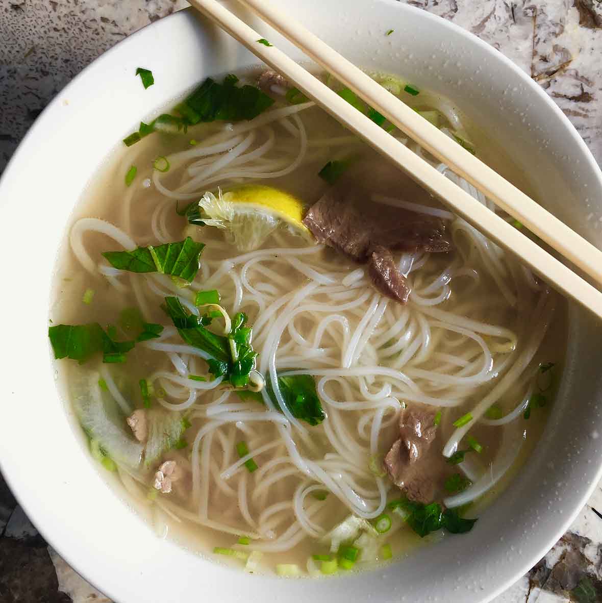 A bowl of authentic Vietnamese pho with rice noodles, beef, lemon, basil, and scallions in it and a pair of chopsticks resting on top of the bowl.