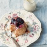 A microwave mug cake upside-down plum cakeon a floral plate beside a glass pitcher of heavy cream.