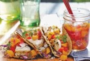 An open jar of peach salsa next to three fish tacos topped with peach salsa.