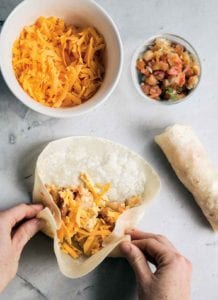 A person assembling a breakfast burrito and a complete breakfast burrito, and bowls of cheese and salsa on the side