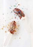 Two chocolate banana pops, one covered in sprinkles and the other in chopped nuts on a piece of parchment.
