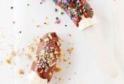 Two chocolate banana pops, one covered in sprinkles and the other in chopped nuts on a piece of parchment.