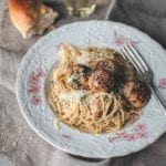 A patterned bowl filled with turkey meatballs with angel hair pasta, and a piece of bread and glass of white wine next to the bowl.