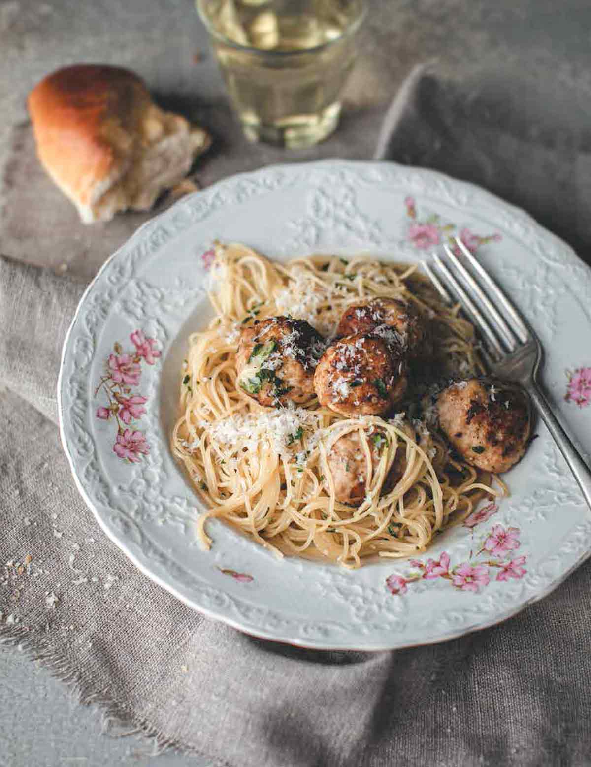 A patterned bowl filled with turkey meatballs with angel hair pasta, and a piece of bread and glass of white wine next to the bowl.