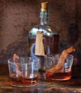 Two glasses of bacon bourbon with strips of cooked bacon in each glass and a bottle of bourbon in the background.