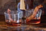 Two glasses of bacon bourbon with strips of cooked bacon in each glass and a bottle of bourbon in the background.