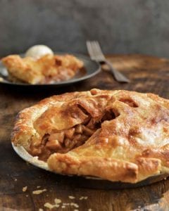 A Northern Spy apple pie in a pie plate with one slice missing, with that piece on a plate in the background.