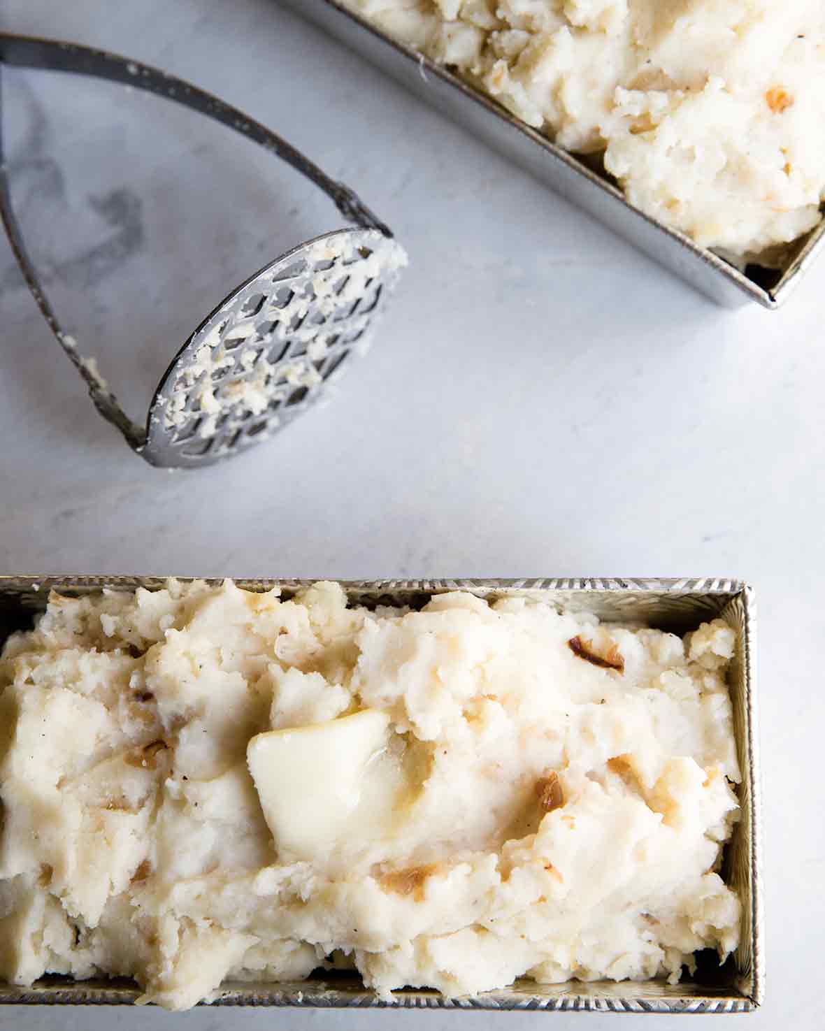 Two metal loaf pans filled with roasted garlic mashed potatoes with a masher resting in between them.