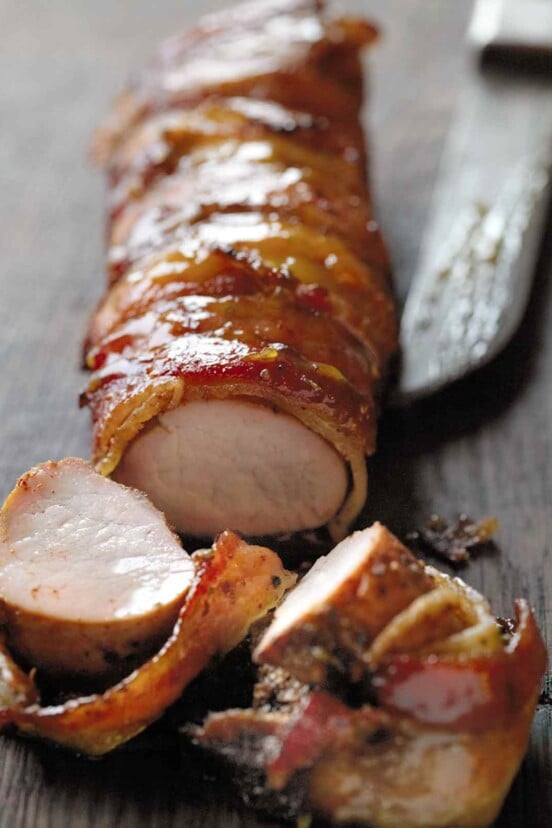 A bacon-wrapped pork tenderloin with a few slices cut from the end and a knife resting beside.