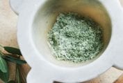 A mortar partially filled with herb salt and a pestle and two sprigs of fresh sage leaves lying beside it.