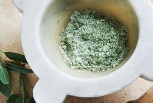 A mortar partially filled with herb salt and a pestle and two sprigs of fresh sage leaves lying beside it.