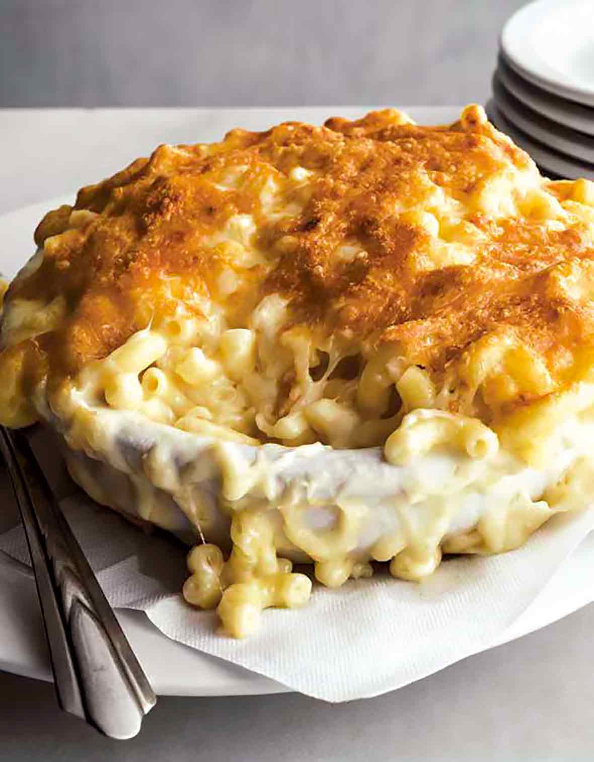 A dish of macaroni au gratin on a white paper napkin on a white plate with a stack of plates beside it.