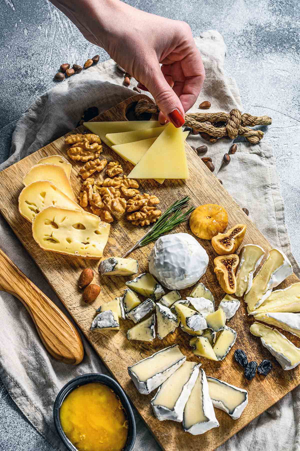Various cheeses and nuts on a wooden cutting board, with a woman's hand picking up a triangle of cheese.
