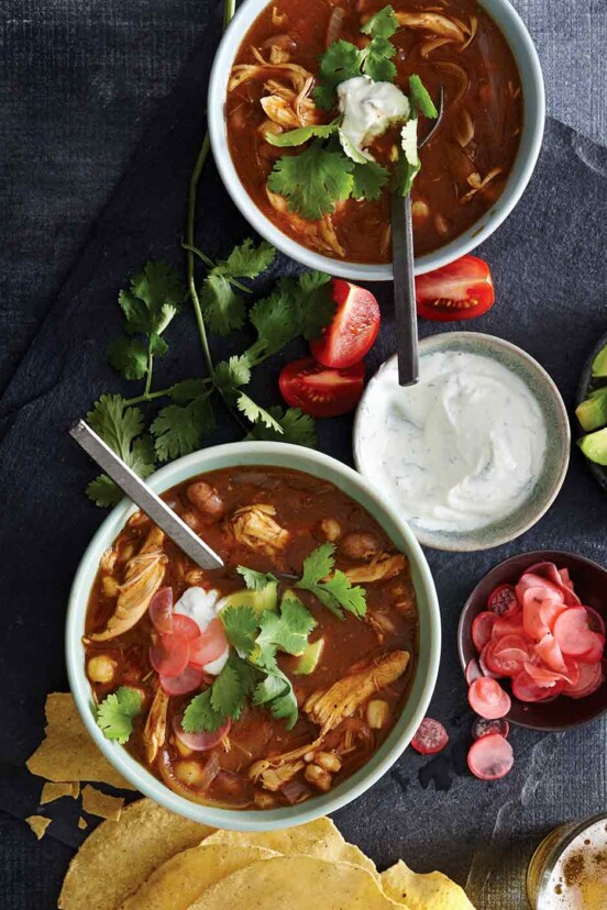 Two bowls of chicken posole, with a bowl of sour cream, some tomato wedges, cilantro, sliced radish, and tortilla shells scattered around them.