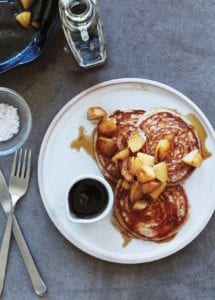 A plate with a small dish of maple syrup and 3 rye pancakes that are topped with maple syrup and pears.