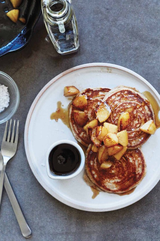 A plate with a small dish of maple syrup and 3 rye pancakes that are topped with maple syrup and pears.