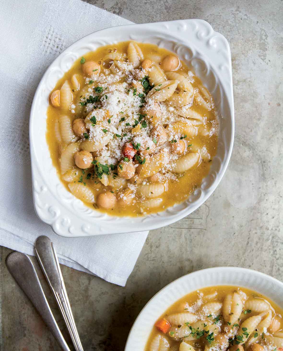 Two white bowls filled with chickpea and cavatelli soup on white napkin with spoons.