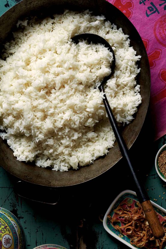 A large wooden bowl of coconut rice with a serving spoon beside assorted garnishes, on a pink tablecloth.