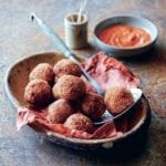 A large bowl filled with fried arancini balls and a small bowl of red pepper sauce on the side