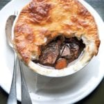 A venison pie with part of the crust missing on a white plate with a fork and spoon beside it.