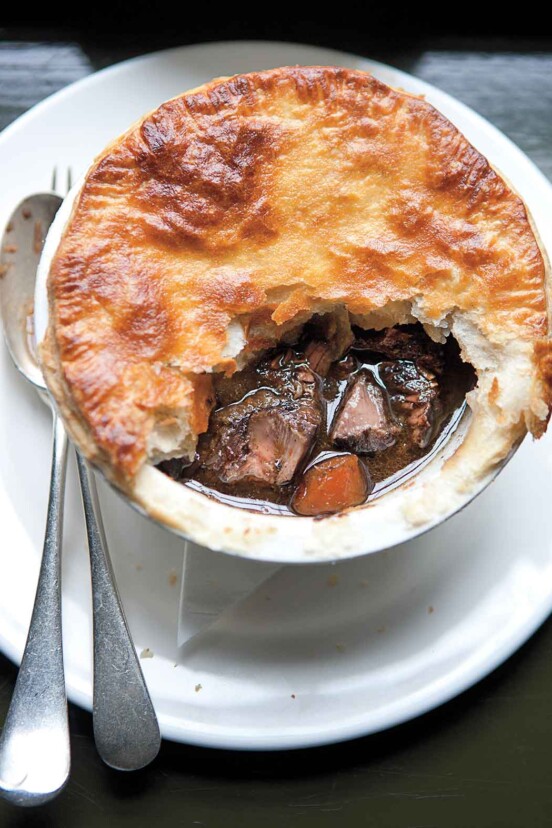 A venison pie with part of the crust missing on a white plate with a fork and spoon beside it.