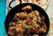 A black pot filled with chunks of beef Raj curry, topped with cilantro, nearby is roti Indian bread