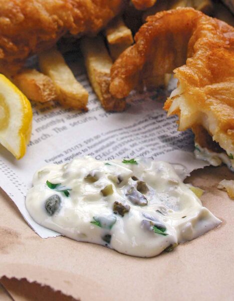 A dollop of homemade tartar sauce on a piece of newspaper with fried fish and a lemon wedge beside it.