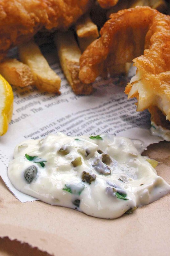 A dollop of homemade tartar sauce on a piece of newspaper with fried fish and a lemon wedge beside it.