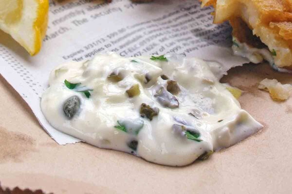 A dollop of homemade tartar sauce on a piece of newspaper with fried fish and a lemon wedge beside it.