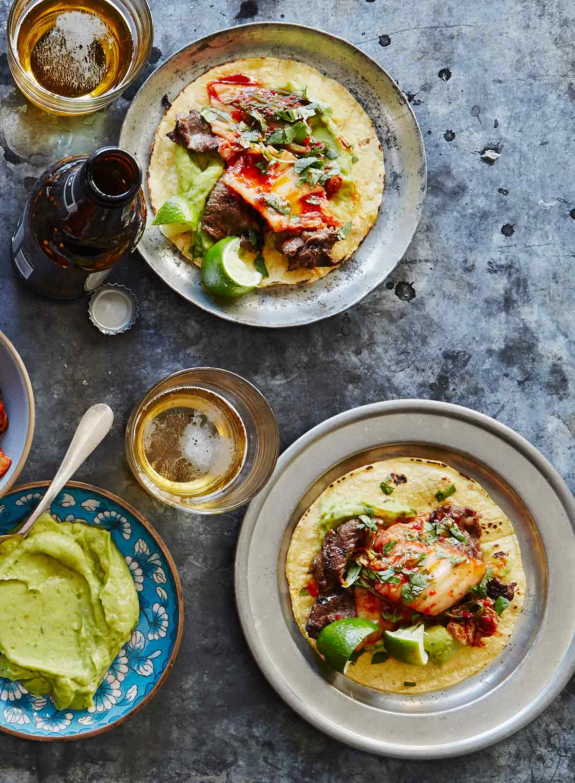 Two plates topped with kimchi tacos with glasses of beer and a bowl of guacamole on the side.