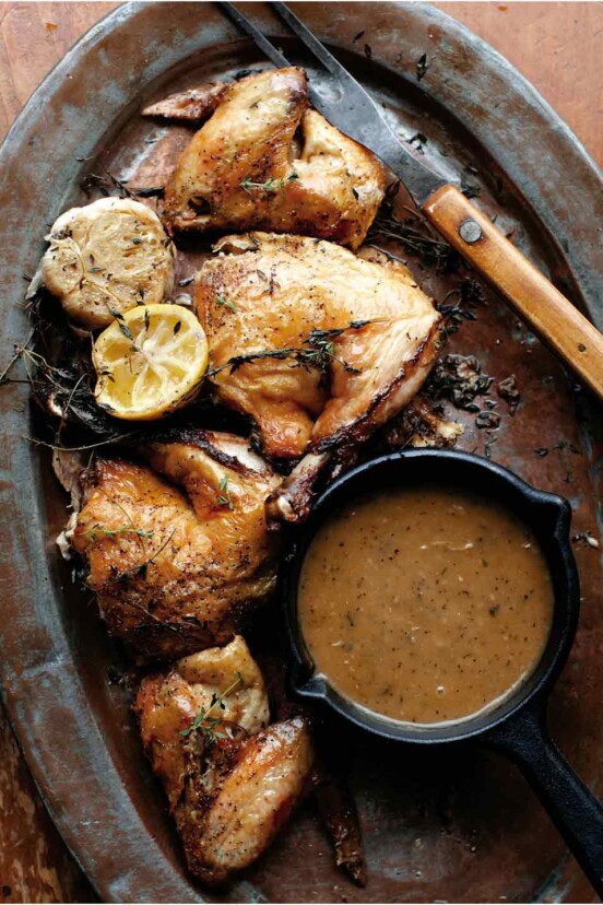 Pieces of roast chicken wth a small dish of pan gravy and a halved lemon and bulb of garlic on an oval metal tray.