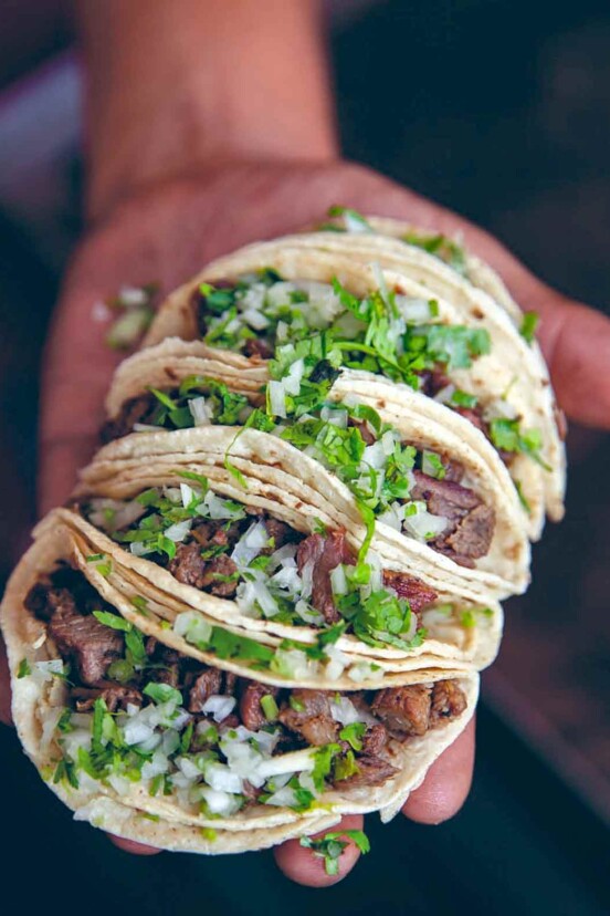 Man's hand holding four beef tongue tacos, or taquitos de lengua, filled with cubed meat, onions, cilantro