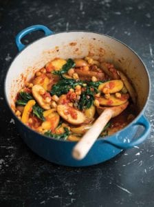 A blue Dutch oven with a potato and chickpea stew and some spinach with a wooden spoon resting inside.