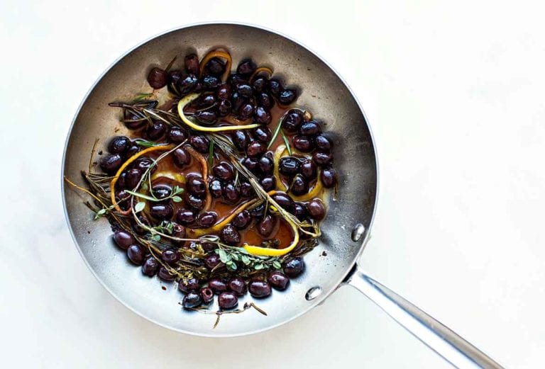 A skillet filled with black olives in an infusion of herbs and lemon and orange peel